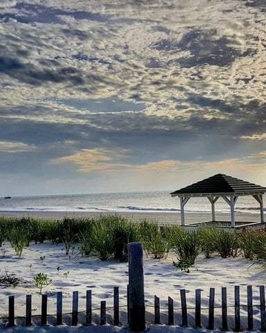 Beach at sunset with wooden gazebo, dune fence, sea grass, and ocean horizon under dramatic cloudy sky