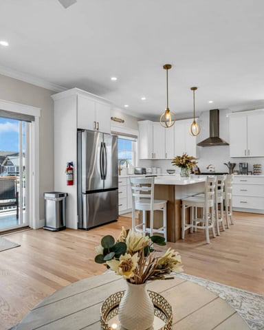 Open-plan kitchen and dining area with white cabinetry, stainless steel appliances, island seating, hardwood floors, and ocean views through glass doors.