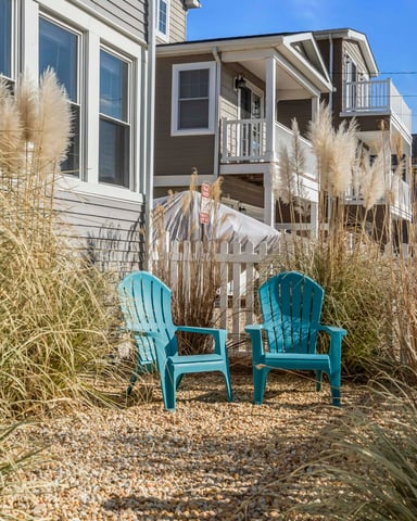 Two turquoise Adirondack chairs on gravel surrounded by dried beach grass, facing beachfront houses under clear blue sky