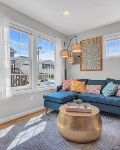 Bright living room with blue sectional sofa, woven pendant lights, round gold coffee table, and large windows overlooking residential neighborhood.