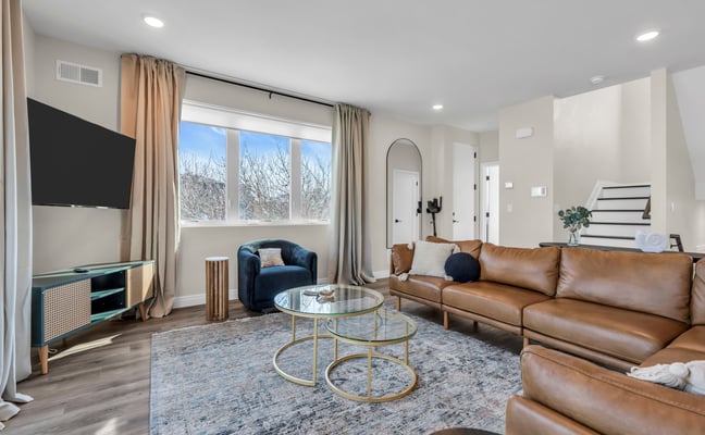 Modern living room with tan leather sectional, blue armchair, glass coffee table, TV, and large windows overlooking bare trees