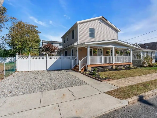 Two-story house with white siding, front porch with white railings, attached garage with lattice gate, gravel driveway, and landscaped yard.