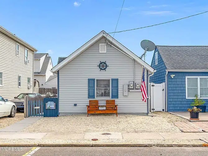 Small white beach cottage with blue shutters, ship wheel decoration, wooden bench, and American flag on sandy lot